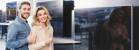 panoramic shot of smiling boyfriend and girlfriend showing thumbs up in home appliance store