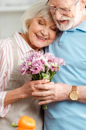 Selective focus of smiling senior couple holding bouquet of chrysanthemums in kitchen