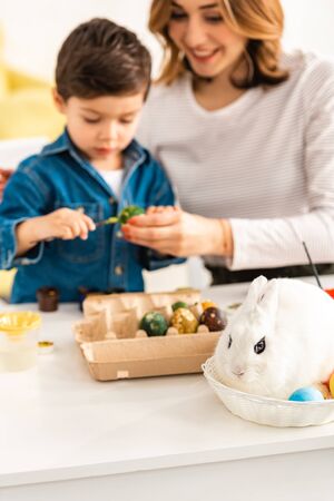 selective focus of mother and son painting Easter eggs while sitting at table near bunny in wickerの写真素材