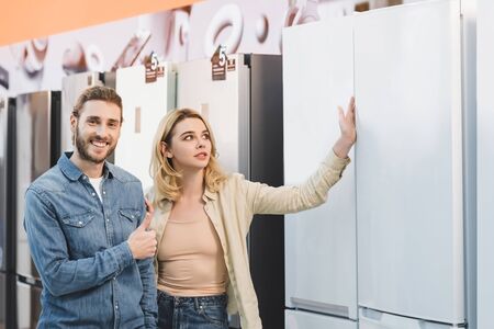 smiling boyfriend showing like and girlfriend touching fridge in home appliance storeの写真素材