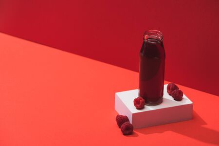 fresh berry juice in glass bottle near ripe raspberries on cube on red backgroundの写真素材