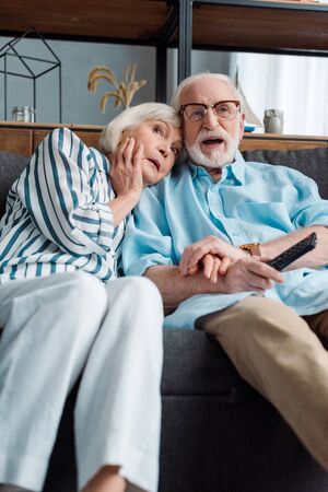 Selective focus of shocked senior couple watching tv on couch at homeの写真素材