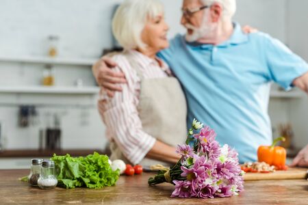 Selective focus of bouquet and fresh vegetables on table and smiling senior couple in kitchenの写真素材