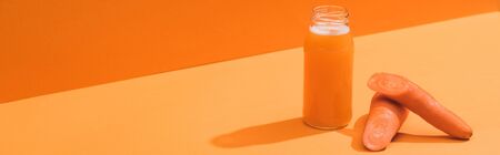 fresh juice in glass bottle near ripe carrots on orange background, panoramic shotの写真素材
