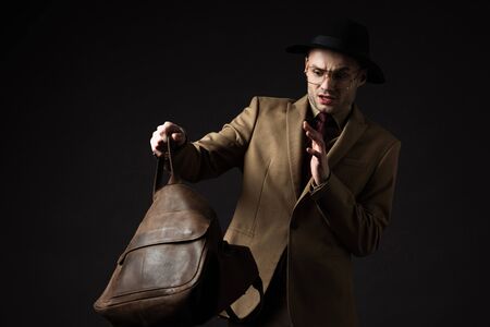 stressed elegant man in beige suit, hat and eyeglasses looking in brown leather bag isolated on...