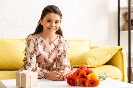 cheerful child in elegant dress looking at camera while sitting near table with flowers and gift box