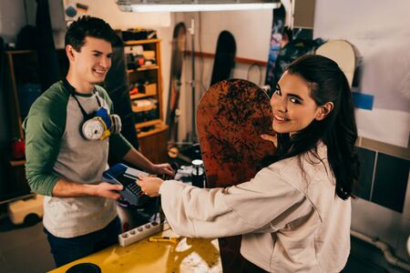 worker holding terminal and smiling woman paying with credit card and holding snowboard in repair...