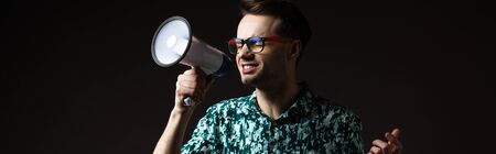 fashionable man in eyeglasses in blue colorful shirt shouting in loudspeaker isolated on black,...