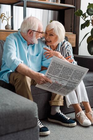 Senior couple smiling at each other and man holding newspaper on couch at homeの写真素材