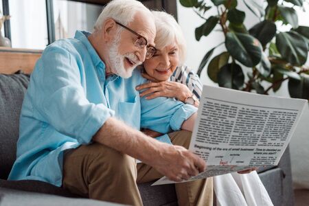 Smiling senior couple reading newspaper on couch in living roomの写真素材