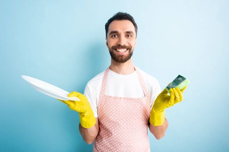 cheerful man in dotted apron holding plate and sponge on blueの写真素材