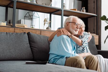 Selective focus of senior couple hugging by remote controller on couch at home
