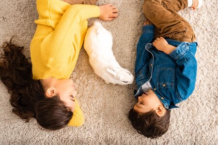 top view of cute brother and sister lying on floor near white funny rabbitの写真素材