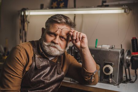 senior, confident cobbler looking at camera while sitting near sewing machine in workshop