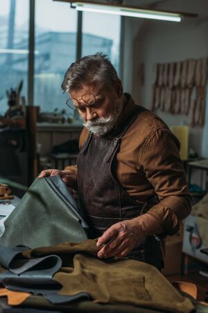 attentive, senior cobbler looking at pieces of genuine leather in workshop