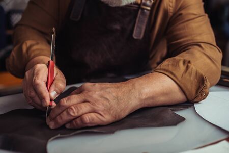 partial view of cobbler cutting genuine leather with knife