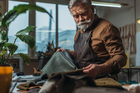 selective focus of senior cobbler holding pieces of genuine leather in workshopの写真素材