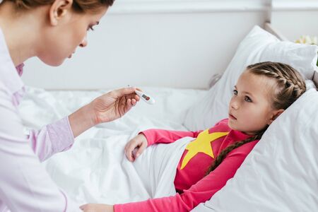worried mother looking at thermometer with sick daughter in bed