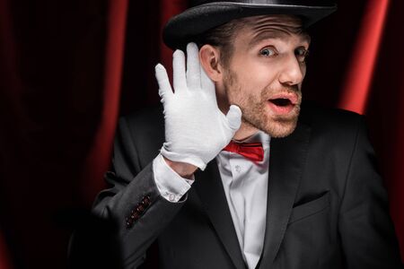 confused magician gesturing and listening in circus with red curtains