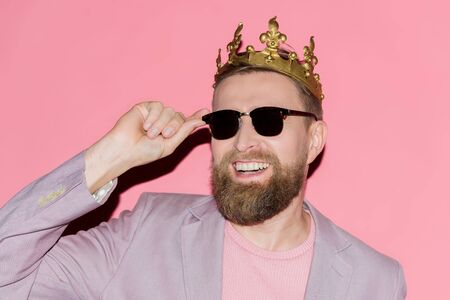 smiling man with crown touching sunglasses on pink backgroundの写真素材