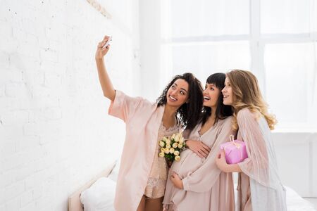 smiling multicultural girlfriends and happy pregnant woman holding flowers and present while taking selfie on baby showerの写真素材