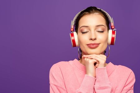 smiling, dreamy girl listening music in wireless headphones with closed eyes isolated on purpleの写真素材