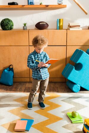 cute and smart kid writing in notebook while standing near books and appleの写真素材