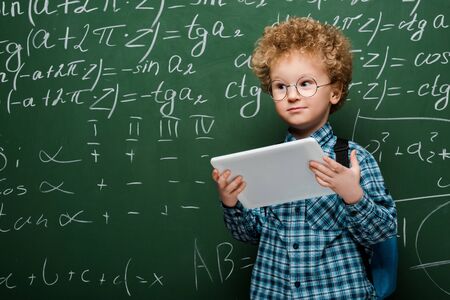 smart kid in glasses holding digital tablet near chalkboard with mathematical formulas