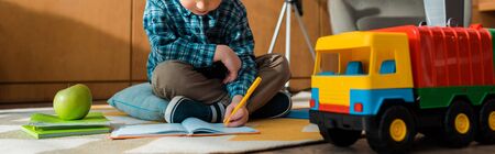 panoramic shot of smart kid writing in notebook near toy car and apple while sitting on floor