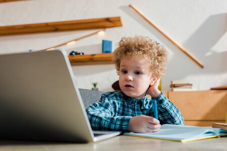 selective focus of kid holding pen while studying near laptop at homeの写真素材