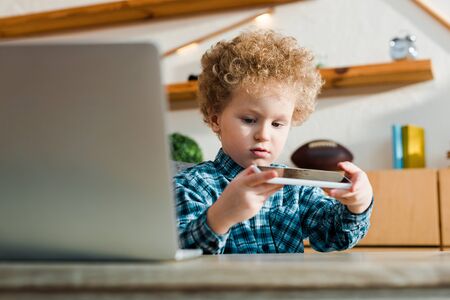 selective focus of smart child looking at smartphone near laptopの写真素材