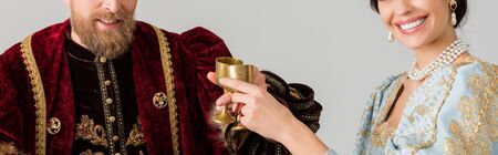 panoramic shot of smiling queen and king clinking with cups isolated on greyの写真素材