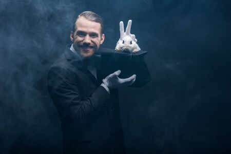 positive magician in suit showing trick with white rabbit in hat, dark room with smokeの写真素材