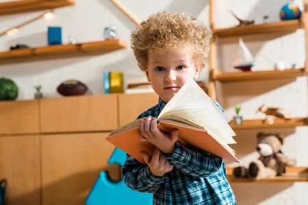curly kid holding book and looking at camera at home