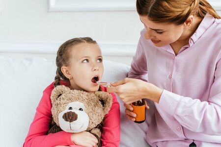 mother giving syrup to ill daughter with teddy bearの写真素材