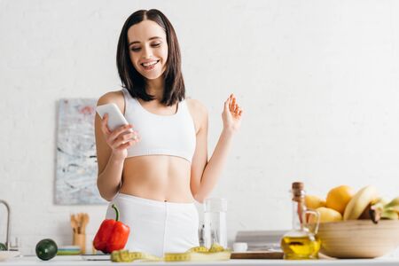 Low angle view of smiling sportswoman using smartphone near fruits, vegetables and measuring tape in kitchenの写真素材
