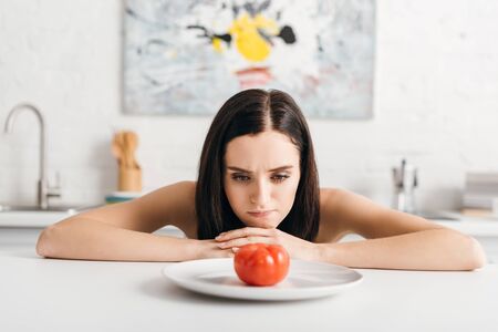 Selective focus of pensive girl looking at ripe tomato on kitchen tableの写真素材
