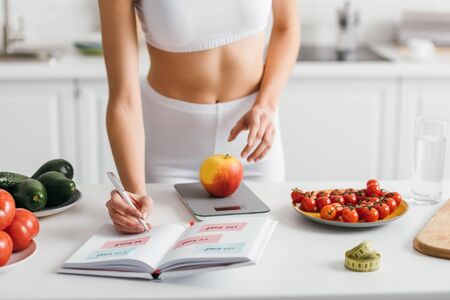 Cropped view of fit sportswoman writing calories while weighing apple on kitchen table, calorie counting dietの写真素材