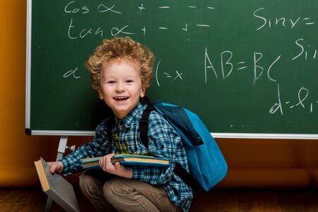 happy kid laughing while holding books near chalkboard with mathematical formulas