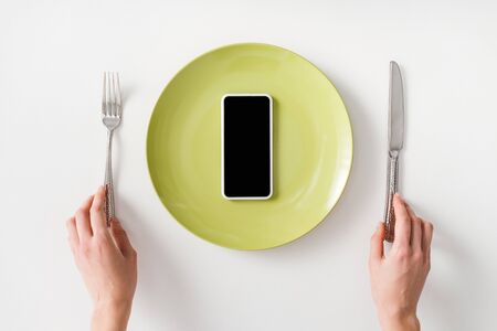 Top view of female hands with cutlery and smartphone on plate on white background
