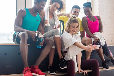 Smiling multicultural  dancers taking selfie while resting on bench in dance studio