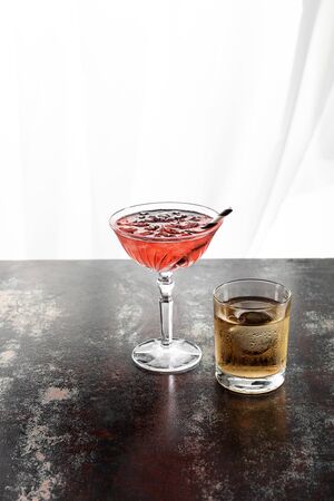 wet glass of whiskey near red cosmopolitan cocktail with ice cubes on whiteの写真素材