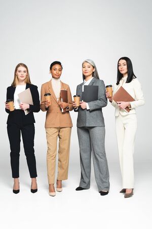 four multicultural businesswomen holding folders and paper cups while standing on whiteの写真素材
