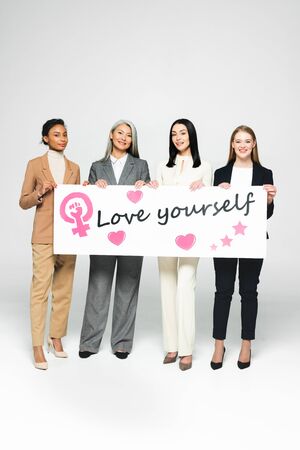 happy multicultural businesswomen holding poster with love yourself lettering on whiteの写真素材