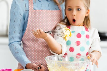 Selective focus of shocked and excited child looking at balloon whisk in dough above bowl