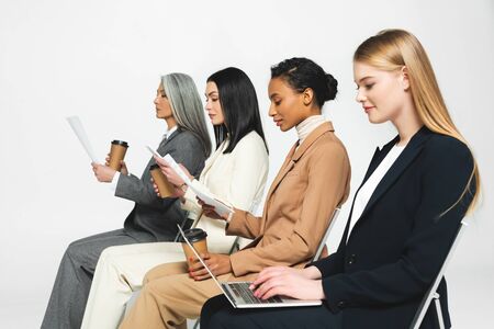 side view of multicultural businesswomen sitting on chairs with paper cups and laptop on whiteの写真素材
