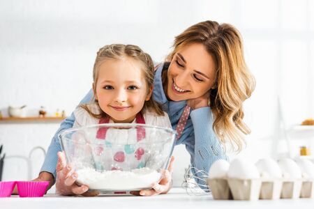 Selective focus of happy mom and cute daughter holding bowl with flour in kitchen