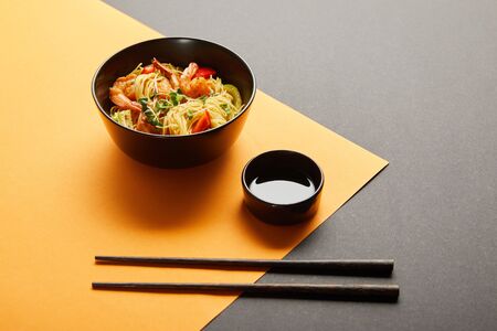noodles with shrimps and vegetables in bowl near chopsticks, soy sauce on black and yellow...