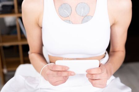 Cropped view of woman holding smartphone during electrotherapy of chest in clinic