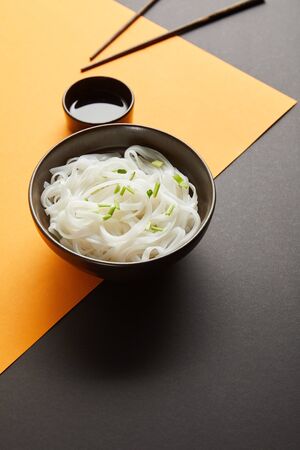rice noodles in bowl near chopsticks and soy sauce on yellow and black surface